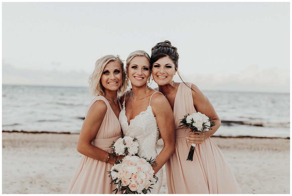 bride and bridesmaids in long blush dresses taking photos on the beach before wedding ceremony in cancun mexico
