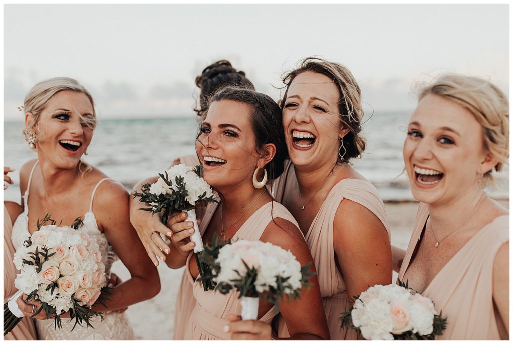 bride and bridesmaids in long blush dresses taking photos on the beach before wedding ceremony in cancun mexico