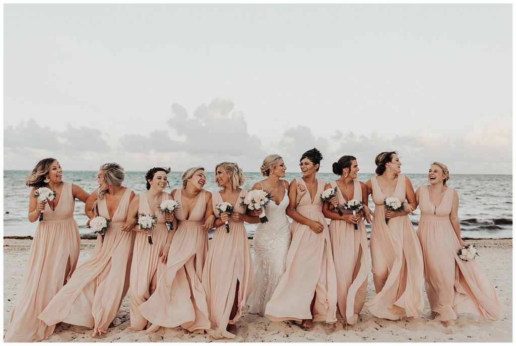 bride and bridesmaids in long blush dresses taking photos on the beach before wedding ceremony in cancun mexico