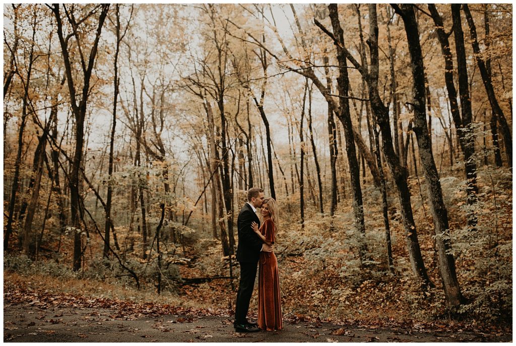 Engaged couple taking portraits in Kentucky, surrounded by fall leaves. Girl is wearing a rust colored velvet dress