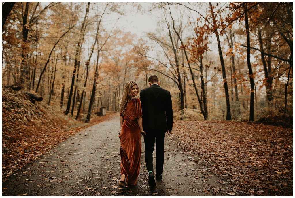Engaged couple taking portraits in Kentucky, surrounded by fall leaves. Girl is wearing a rust colored velvet dress