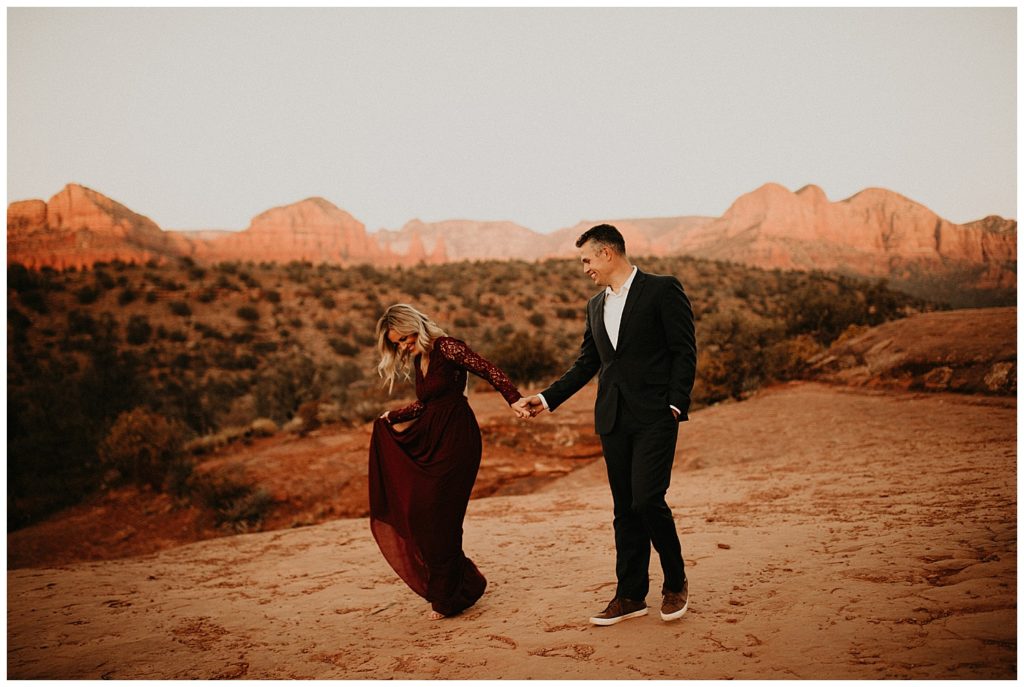 engaged couple holding hands while  taking portraits in sedona, girl is wearing long red velvet dress